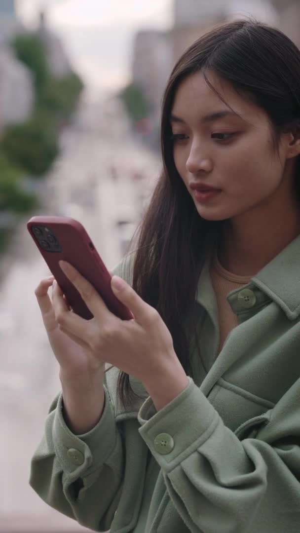 Angenehme Frau in Freizeitkleidung, die auf den Telefonbildschirm schaut und lächelt, während sie auf der Straße der Stadt steht. Asiatische Brünette SMS auf modernen Smartphone im Freien. Vertikales Video — Stockvideo