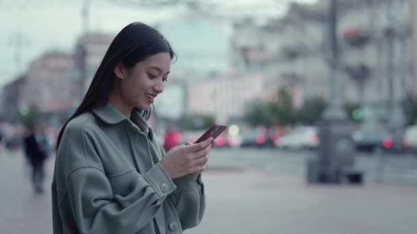 옥외에서 아시아인 여자가 스마트폰으로 문자를 보내는 휴대용 화면을 보면서 미소짓는 — 비디오