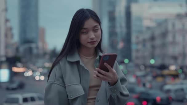 Ritratto di bella donna asiatica che messaggia su smartphone e guarda la macchina fotografica mentre si trova in strada durante l'ora di sera. Concetto di giovani, gadget e stili di vita moderni — Video Stock