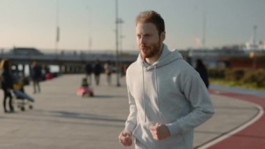 Sakallı, spor kıyafetli genç adam şehir caddesinde koşuyor.