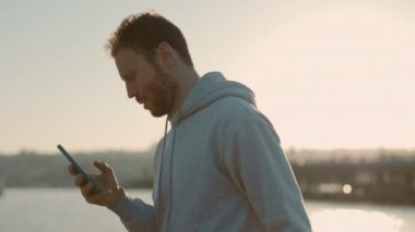 Kızıl saçlı adam şehir nehri yakınlarında koşarken akıllı telefon kullanıyor.