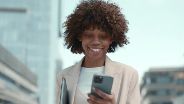 Улыбающаяся черная леди идет по улице и смотрит на мобильный телефон — стоковое видео
