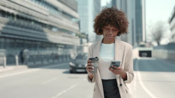 Черная бизнес-леди ходит по улице с кофе и мобильным телефоном — стоковое видео