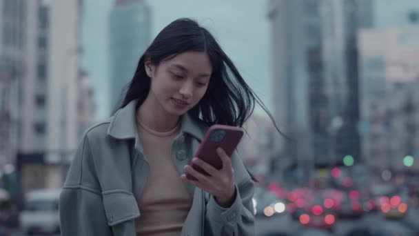 Ritratto di affascinante giovane donna che legge alcune buone notizie su smartphone che tiene in mano. Bruna asiatica utilizzando il telefono cellulare sulla strada della grande città — Video Stock