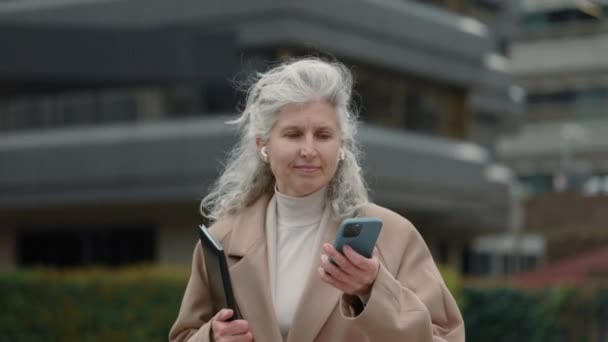 Femme mûre dans les écouteurs marchant et utilisant un téléphone portable — Video