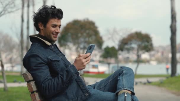 Vista lateral del hombre relajado sentado en el banco y utilizando el móvil — Vídeo de stock