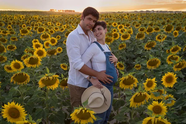 Cheerful Caucasian couple on maternity shoot in a sunflower field during sunset