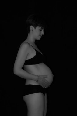 İç çamaşırı giyen ve karnını siyah bir arka planda tutan hamile bir kadın.