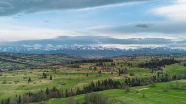 Güney Polonya 'nın kırsal kesimlerinde ilerleyen ve Tatra Dağları' nın karla kaplı zirvelerine doğru ilerleyen hava hiperhız videosu. Yeşil yay tarlaları, dağılmış ağaçlar, dinamik bir gökyüzünün altında uzak dağlık tepeler..