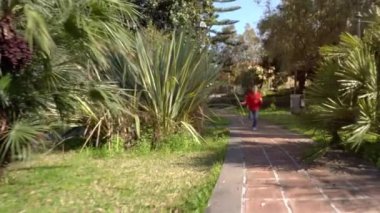 Kırmızı tişörtlü 6 yaşındaki bir çocuk parkta tek başına mutlu bir şekilde koşuyor.