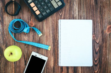 Fitness, sağlıklı ve aktif yaşam tarzı konsepti. Spor ayakkabıları, akıllı telefon, bant, hesap makinesi, spor bilekliği, yeşil elma ve tahta arka plandaki defter.