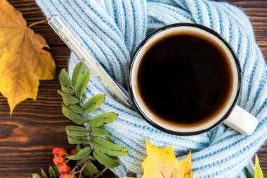 Termometresi, mavi eşarbı ve ahşap arka planda sonbahar yaprakları olan çay fincanı. Sonbaharda grip mevsimi, hastalık.