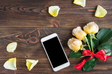 Kırmızı fiyonklu sarı güller, tebrik kartı ve ahşap arka planda cep telefonu. Kadınlar Günü. Sevgililer Günü. Doğum günü.