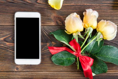 Kırmızı fiyonklu sarı güller, tebrik kartı ve ahşap arka planda cep telefonu. Kadınlar Günü. Sevgililer Günü. Doğum günü.