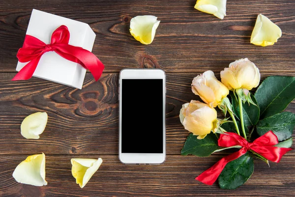 Yellow roses with red bow, greeting card and mobile phone on wooden background. Women\'s day. Valentines day. Birthday.