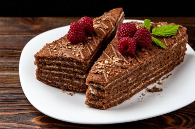 Slice of chocolate cake with milk filling and raspberry on white plate on dark wooden background.