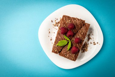 Slice of chocolate cake with milk filling and raspberry on white plate on blue background.