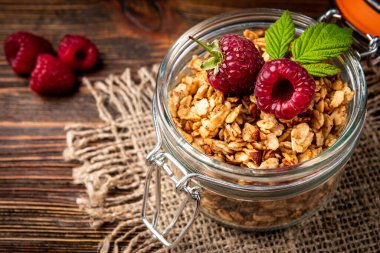 Granola in glass jar and raspberry on dark wooden background.