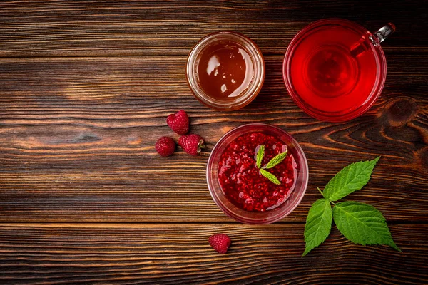 Cup of raspberry tea with raspberry jam and honey on dark wooden background.