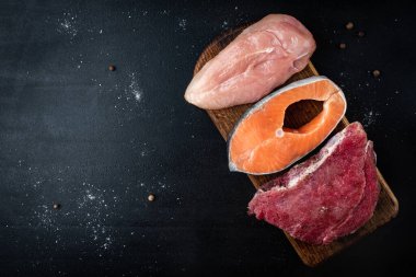 Taze çiğ et, somon balığı ve tavuklu fileto. Siyah arka planda ahşap kesim tahtası. Yüksek proteinli doğal besinler. Üst görünüm.
