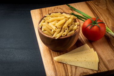 Tahta kasede çiğ makarna, peynirli, domatesli ve arka planda yeşil soğanlı..