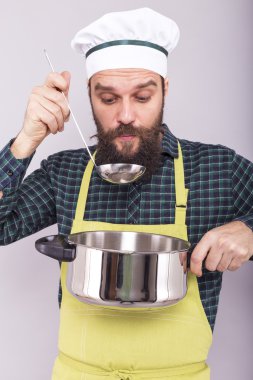 Stüdyo resmi, mutlu bir aşçının gri arka planda kepçeyle çorba tatması.