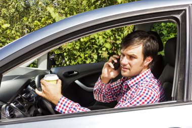  Young man speaking on phone and drinking coffee while driving  clipart