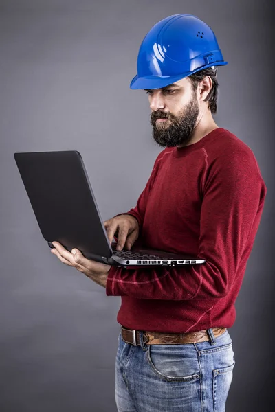 Junger Ingenieur mit Hut steht während er an seinem Laptop arbeitet — Stockfoto