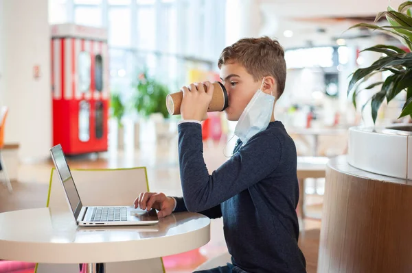 Ensino à distância. Um adolescente em uma máscara médica senta-se a uma mesa em um café com um laptop e uma caneca de bebida quente.. — Fotografia de Stock