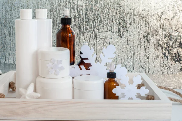 A set of winter care cosmetics against the background of the winter window with frosty patterns. White jars and vials of drips on a white table. — Stock Image A set of winter care cosmetics against the background of the winter window with frosty patterns. White jars and vials of drips on a white table. Royalty Free Stock Photos