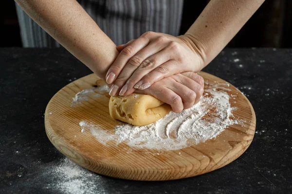 O processo de cozinhar a massa. As mãos das mulheres amassam a massa em uma tábua redonda de madeira em uma mesa escura. — Fotografia de Stock
