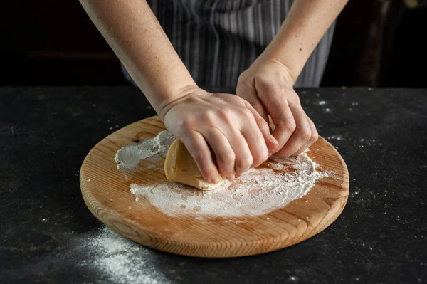 ペストリーを調理するプロセス。女性の手は暗いテーブルの上に木製の丸板の上に生地をこねる. — ストック写真