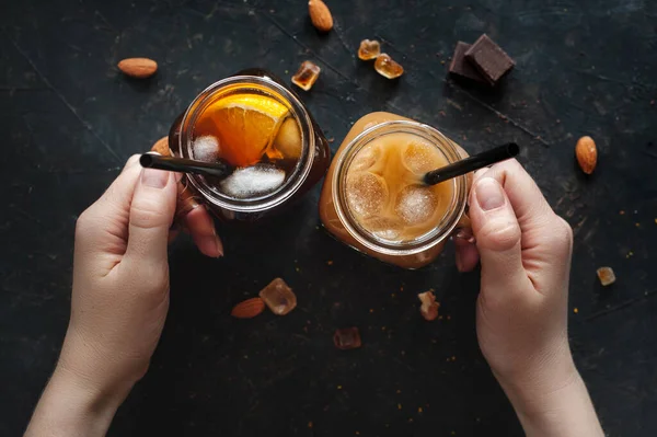 Cold espresso with ice and orange juice in two cans in womens hands above a dark table. — Stock Photo, Image