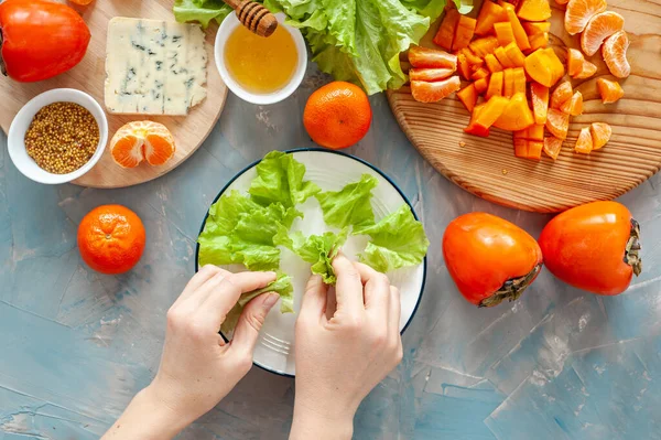 Ingredientes y el proceso de hacer una ensalada de vitamina invierno con caquis, mandarinas y queso azul. Paso a paso. La mujer pone las hojas de lechuga en un plato. Vista desde arriba. — Foto de Stock