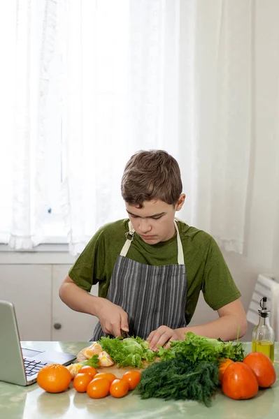 Мальчик-подросток готовит фруктовый салат по рецепту из интернета. Мальчик режет салат на кухонном столе, перед ним ноутбук. Ребенок занимается домашними делами. — стоковое фото