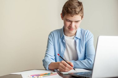 Onlay bir çizim dersi. Genç bir çocuk bir albümde kalem çiziyor, önünde kalemler ve dizüstü bilgisayarlar var..