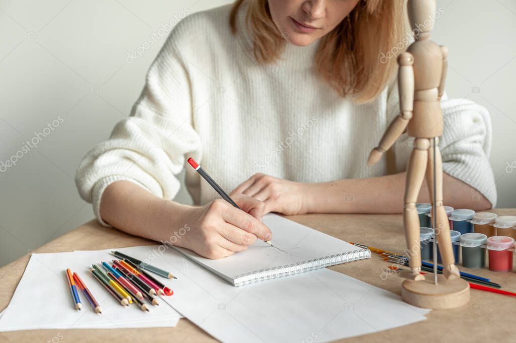 A young beautiful blonde woman in a white jumper sits at the table and draws a man on the model. Drawing. Hobby. Mental health. A place for text.