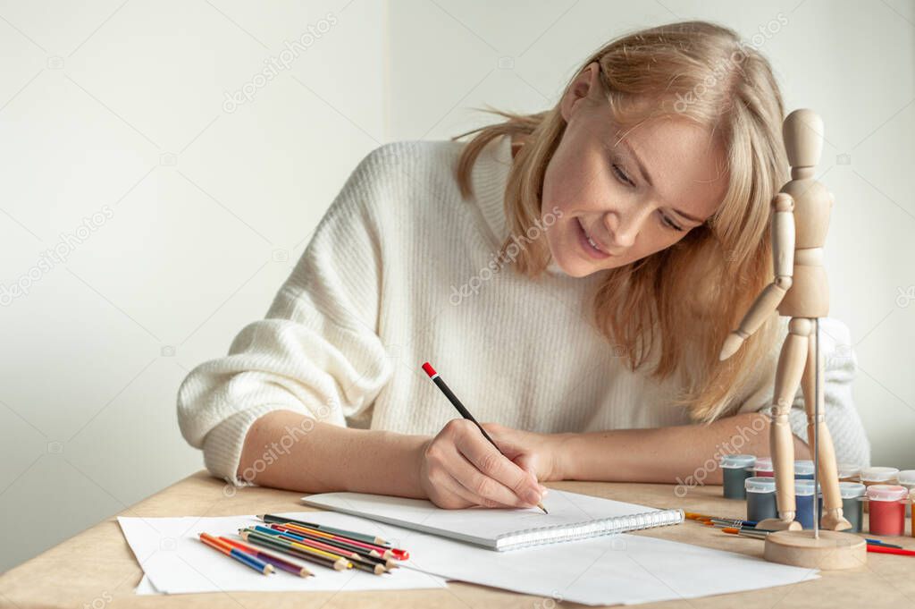 A young beautiful blonde woman in a white jumper sits at the table and draws a man on the model. Drawing. Hobby. Mental health. A place for text.