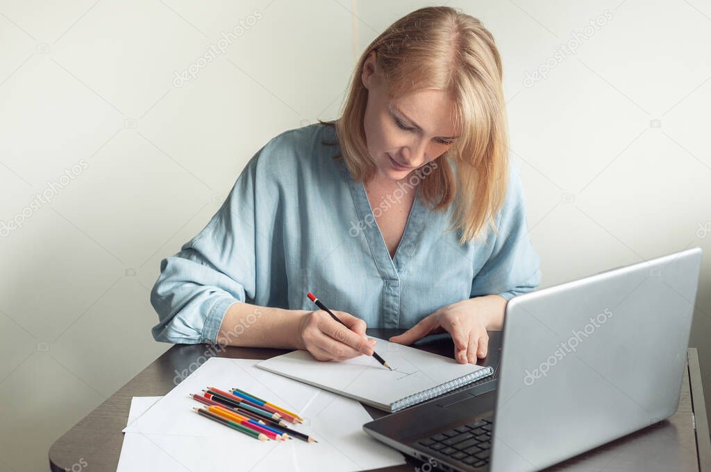 Hobbies and online training. A beautiful blonde woman draws in an album on an online lesson. A woman draws with a pencil in front of a laptop.