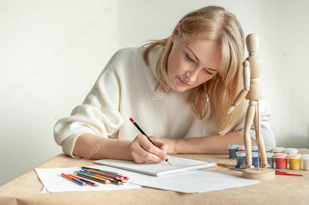 A young beautiful blonde woman in a white jumper sits at the table and draws a man on the model. Drawing. Hobby. Mental health. A place for text.