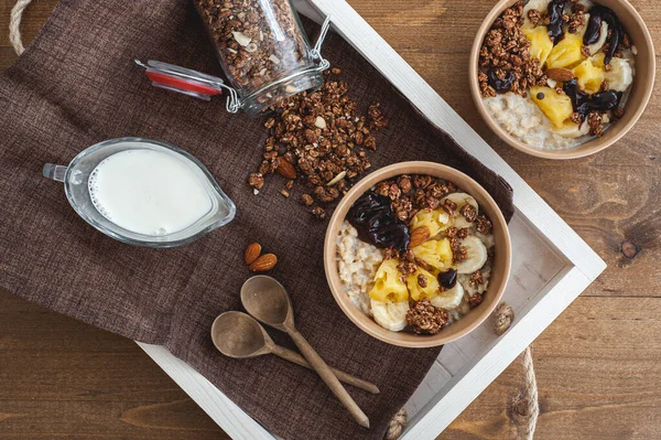 Farina d'avena con ananas, banana e granola di cioccolato in piatti beige su un tavolo di legno marrone. Una ricca colazione sana. Un posto per il testo. — Foto Stock