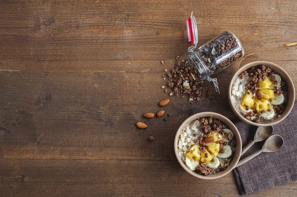 Harina de avena con piña, plátano y granola de chocolate en platos beige sobre una mesa de madera marrón. Un desayuno saludable. Un lugar para el texto. —  Fotos de Stock