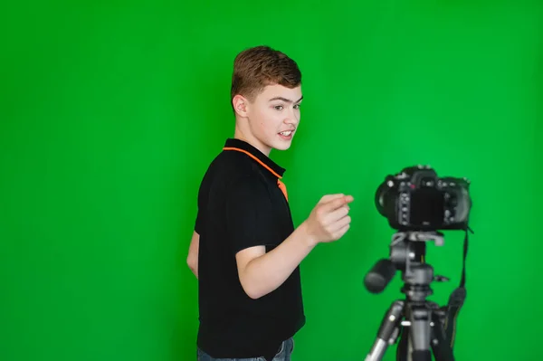 Un bloguero adolescente niño dispara un video en cámara sobre un fondo verde. Una adolescente caucásica con una camiseta negra emocionalmente dice algo para un blog. —  Fotos de Stock