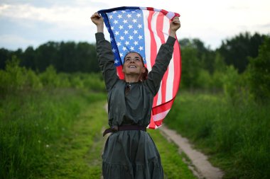 Uzun saçlı, genç, mutlu bir Amerikalı kadın, rüzgarda dalgalanan Amerikan bayrağını omuzlarında tutuyor, açık havada dinleniyor, sıcak bir yaz gününün tadını çıkarıyor. 4 Temmuz