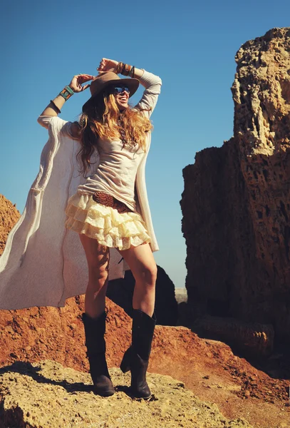 Foto de moda al aire libre de la joven mujer feliz en el sombrero, sentado en una piel contra las rocas, manos arriba —  Fotos de Stock