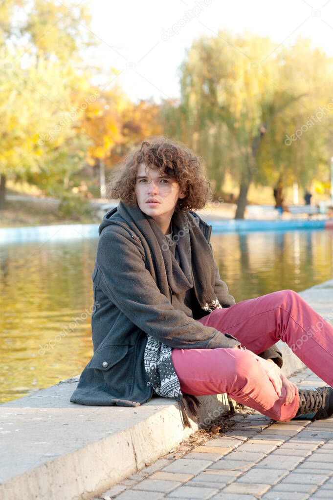 Young pensive teen man portrait sitting near the city lake oudoor in autumn park