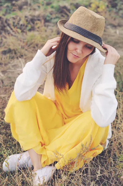 Vrouw gekleed in gele jurk, witte trui en hoed zittend op herfst veld. — Stockfoto