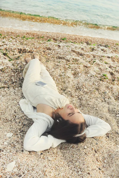 Female dressed in white clothing lying on a sea sand and listening music. — Stock Photo, Image