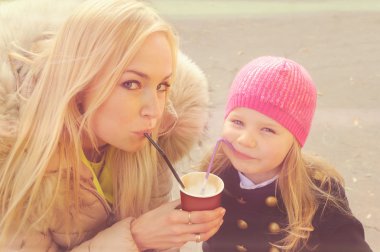Cute young mother with daughter have a fun drinking a milkshake from the same paper cup.