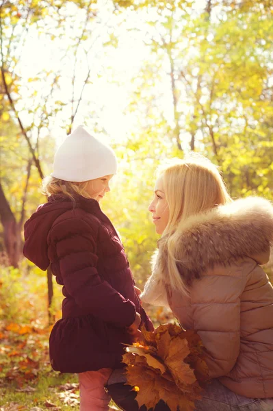 Small daughter and her mother looking at each other and smiling, happy childhood, backlight in autumn park. — Stok fotoğraf